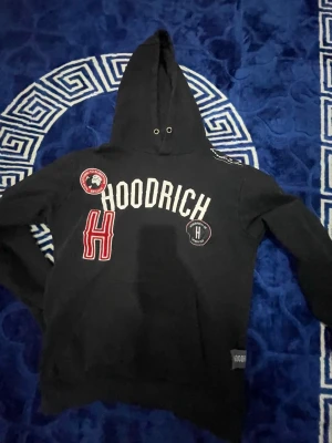 Svart hoodie från Hoodrich - Säljer en svart hoodie från Hoodrich med stora vita och röda tryck och patches på bröstet. Hoodien har en klassisk huva med dragsko och långa ärmar. Perfekt för dig som gillar streetwear och vill sticka ut med snygga detaljer.