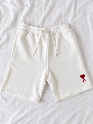 Vita shorts med snörning och hjärtdetalj - Säljer ett par vita shorts med snörning i midjan och en röd hjärtdetalj med bokstaven A på vänster ben. Shortsen har elastisk midja och är tillverkade i mjukt material, perfekta för sommardagar.