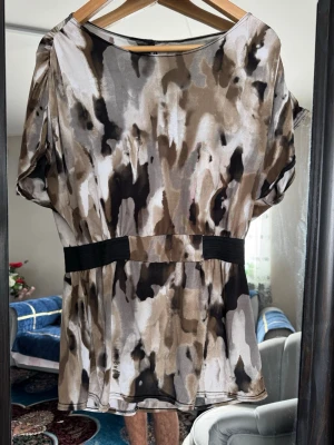 Kort klänning med abstrakt mönster - Kort klänning i mjukt, glansigt material med abstrakt mönster i beige, svart och brunt. Modellen har korta ärmar med lätt puff och markerad midja med svart band. Perfekt för dig som gillar unika prints och avslappnad passform.