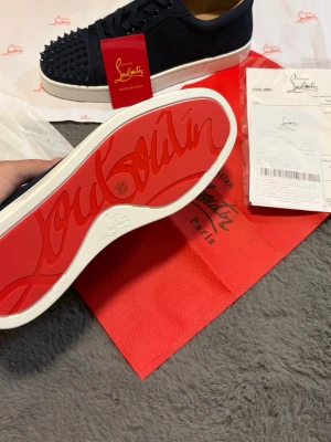 Svarta Louboutin sneakers med nitar - Snygga svarta Christian Louboutin med klassisk röd sula och svarta nitar på tån. Skorna har en låg profil, vita sulor. Perfekta för dig som vill sticka ut med en edgy och exklusiv look.
