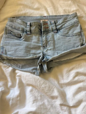 Ljusblå denimshorts med broderi - Säljer ett par ljusblå denimshorts med dubbla knappar och fickor fram och bak. På bakfickorna finns ett coolt broderat mönster i rött och vitt. Shortsen har rå kant nertill för en avslappnad look och bälteshällor i midjan.