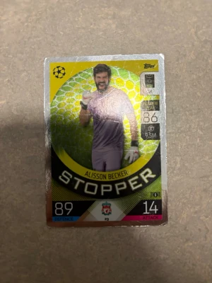 Match Attax Alisson Becker kort - Samlarbild från Match Attax med Alisson Becker som målvakt för Liverpool FC. Kortet har gul och svart design med texten 'STOPPER' och visar spelarens statistik. Perfekt för dig som gillar fotboll och samlarkort.