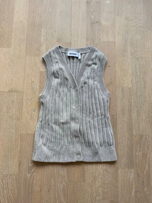 Beige ribbad stickad väst - Snygg beige stickad väst med ribbad struktur och v-ringning. Västen har knappar framtill och är ärmlös, perfekt att styla över en skjorta eller t-shirt för en trendig lager-på-lager-look.
