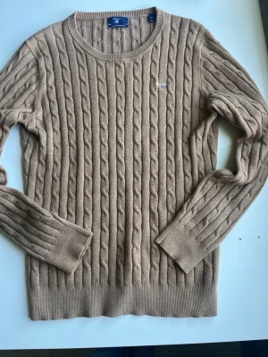 Beige kabelstickad tröja från Gant - Snygg beige kabelstickad tröja från Gant med rund halsringning och diskret logga på bröstet. Tröjan har långa ärmar och ribbade muddar vid ärmslut och nederkant. Perfekt för lager på lager och en klassisk look.