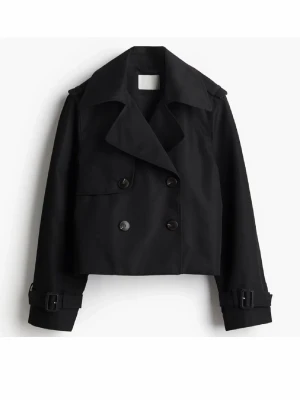 Kort svart trenchcoat med bältdetaljer - Stilren svart trenchcoat i kort modell med dubbelknäppning och bred krage. Jackan har bältdetaljer vid ärmsluten och en ficka framtill. Perfekt för dig som gillar en modern och edgy look. Sparsamt använd!