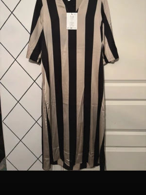 Randig långklänning med v-ringning - Snygg långklänning med breda vertikala svarta och beige ränder. Klänningen har v-ringad hals och trekvartsärm. Perfekt för dig som gillar en stilren och trendig look.