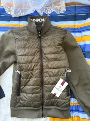 Olivgrön cardiganjacka från Moncler - Snygg olivgrön cardiganjacka från Moncler med quiltad framsida och stickade ärmar. Jackan har hög krage med Moncler-logga, två dragkedjefickor och hel dragkedja framtill. Materialmix av glansig nylon och stickad bomull ger en modern look.
