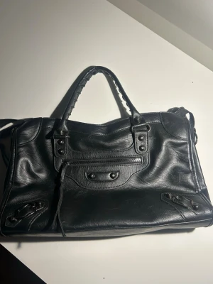 Svart handväska i läderlook - Snygg svart handväska med klassisk design och detaljer som dragkedja framtill, dekorativa nitar och flätat handtag. Väskan har en mjuk, lite glansig läderlook och passar perfekt till en trendig streetstyle. Praktisk storlek för vardagens essentials.