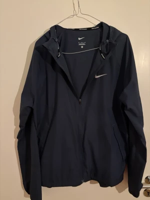 Mörkblå vindjacka från Nike - Snygg mörkblå vindjacka från Nike med huva och dragkedja framtill. Jackan har två sidofickor och en diskret Nike-logga på bröstet. Tillverkad i lätt och vindtätt material, perfekt för blåsiga dagar.