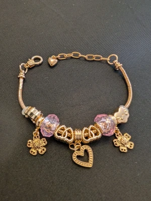 Guldigt armband med pärlor och berlocker - Snyggt armband i guldfärg med lila pärlor och flera detaljerade berlocker, bland annat hjärta och blommor. Armbandet har en kedja med hjärtformad detalj och passar perfekt för dig som gillar unika accessoarer med lite extra bling.