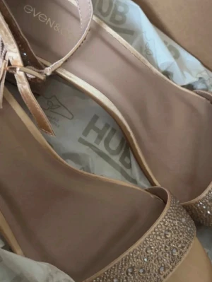 Beige pumps med strassdetaljer - Säljer ett par eleganta beige pumps från even&odd med öppen tå och glittrande strassdetaljer framtill. Skorna har en smal rem runt ankeln och en klassisk siluett som ger en stilren look.