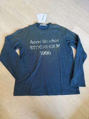 Blå långärmad topp Acne Studios S - Mörkblå långärmad topp från Acne Studios med tryck 'Acne Studios STOCKHOLM 1996' framtill. Klassisk passform och rund halsringning. Perfekt för dig som gillar stilrena och trendiga plagg.