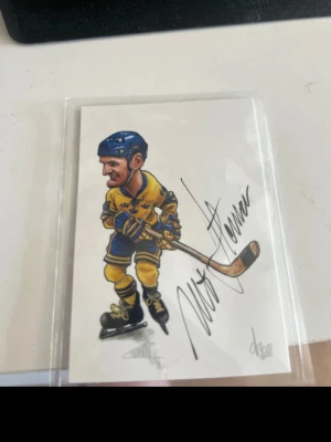 Ishockeykort med tecknad spelare - Unikt samlarkort med tecknad ishockeyspelare i gul och blå matchdräkt, blå hjälm och skridskor. Kortet är signerat och ligger i plastficka. Perfekt för dig som gillar hockey och samlarprylar.