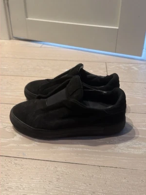 Svarta sneakers från Axel Arigato - Säljer ett par helsvarta sneakers från Axel Arigato i mocka med minimalistisk design och låg profil. Skorna har en diskret look utan synliga snören och en svart gummisula med tydligt mönster. Perfekta för dig som gillar stilrena och cleana skor.