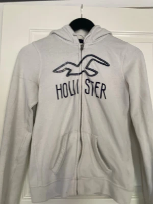 Vit zip hoodie från Hollister - Vit hoodie med dragkedja från Hollister. Stor logga med paljettdetaljer och broderad text på bröstet. Klassisk huva och två fickor framtill. Perfekt för en avslappnad och trendig stil.