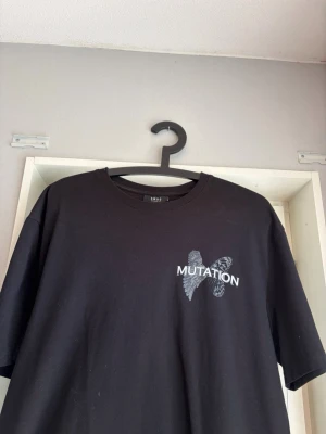 Svart t-shirt med MUTATION-tryck - Cool svart t-shirt med trycket 'MUTATION' och en unik design med två olika vingar på bröstet. T-shirten har rund hals och korta ärmar. Perfekt för dig som gillar streetwear och vill sticka ut lite extra.
