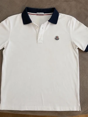 Moncler pikétröja vit - Säljer en Moncler pikétröja i storlek S. Vit med marinblå krage och ärmslut. Mycket fint skick, utan fläckar eller hål. Passar perfekt till både vardag och finare tillfällen.