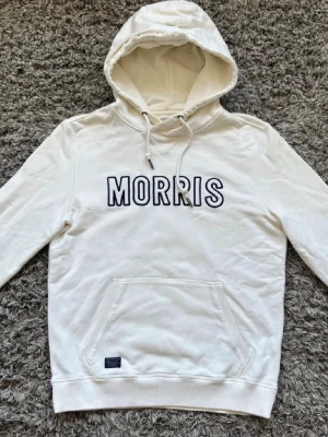 Morris hoodie - Snygg vit hoodie från Morris med svart broderad logga på bröstet. Klassisk känguruficka framtill och justerbar huva med dragsko. Tillverkad i mjuk bomull för skön känsla. Perfekt för en avslappnad och trendig stil.