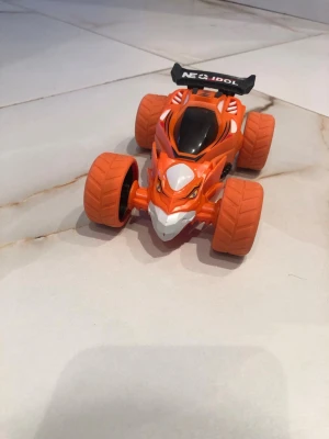 Orange leksaksbil Neo Idol - Cool leksaksbil i plast med stora orange hjul och svart vinge där det står 'Neo Idol'. Bilen har en sportig design med detaljer i vitt och svart samt ett ansikte framtill som ger den en tuff look. Perfekt för dig som gillar fartfyllda leksaker.