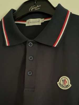 Mörkblå pikétröja från Moncler - Svart pikétröja från Moncler med klassisk krage och knappar framtill. Tröjan har röda och vita ränder på krage och ärmslut samt Moncler-logga på bröstet. Perfekt för en stilren och sportig look.