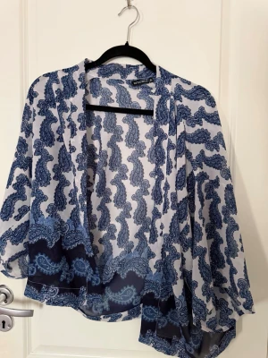 Blåmönstrad blus från Rut&Circle - Säljer en luftig blus från Rut&Circle i storlek 36. Blusen är vit med blått paisleymönster och har långa, vida ärmar. Materialet är tunt och transparent, perfekt för lager på lager. Öppen framtill utan knäppning.