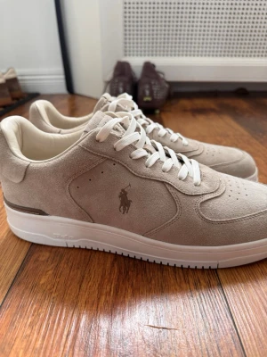 Beige sneakers från Polo Ralph Lauren - Snygga beige sneakers i mocka/beige från Polo Ralph Lauren med klassisk logga på sidan och broderad text på hälen. Skorna har vita snören och en vit sula med diskreta detaljer. Helt nya, bara testade. Köpta från Zalando. Nypris låg på 1800. Digitalt kvitto finns.