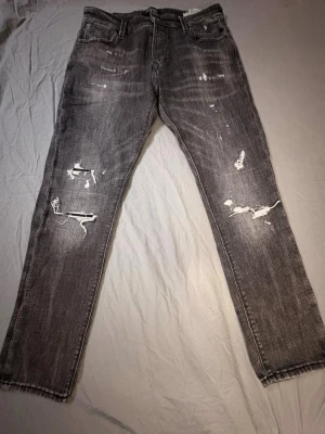 Grå slitna jeans med hål - Säljer ett par grå jeans med trendiga slitningar och hål på båda benen. Jeansen har fem fickor, normal midja och raka ben. Perfekta för dig som gillar en avslappnad stil men åndå vill sticka ut i mängden. Dom är från Jack and Jones och säljs inte längre. 