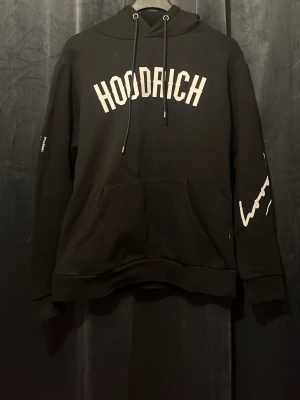 Svart hoodie från Hoodrich - Svart hoodie med vit HOODRICH-text på bröstet och tryck på ryggen. Har huva med dragsnören, stor magficka och tryck längs ena ärmen. Perfekt för en streetwear-look och chill vardagsstil.