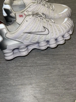 Nike Shox vita sneakers - Nike Shox sneakers i vitt med silverdetaljer och ikonisk fjädrande sula. Ovandelen är i mesh och syntetmaterial för bra andningsförmåga och komfort. Swoosh-logga på sidan och Shox-dämpning ger en futuristisk look. Perfekta för dig som gillar sportig och trendig stil.