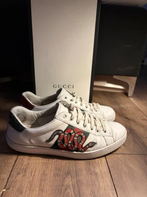 Gucci Ace  - Säljer ett par vita Gucci Ace sneakers i läder med den ikoniska broderade ormen i rött, svart och vitt på sidan. Skorna har gröna och röda detaljer på hälen och klassisk låg silhuett. Perfekta för dig som vill sticka ut med lyxig streetstyle. Storlek 42