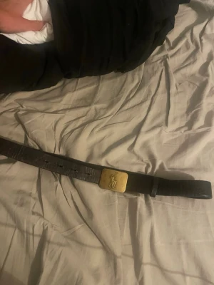 Brunt bälte med guldspänne - Snyggt brunt bälte i skinn med ett fyrkantigt guldspänne som har en ingraverad logga. Perfekt accessoar för att lyfta din outfit och ge en klassisk touch.
