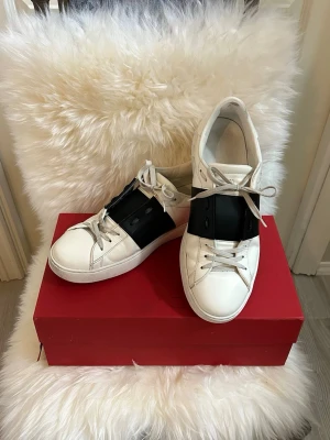 Valentino Garavani  - Snygga vita sneakers från Valentino Garavani med bred svart rem över vristen och vita skosnören. Klassisk låg modell med rund tå och stilren design. Kommer med originalpåse, extra skosnören och äkthetsintyg.