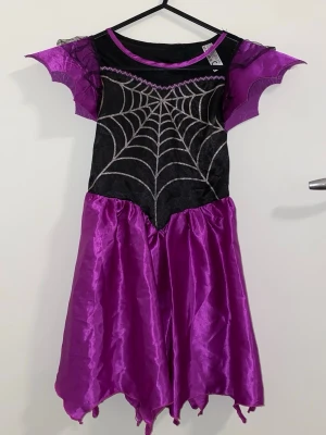 Lila och svart spindelklänning - Cool maskeradklänning i lila och svart med glittrigt spindelnätsmönster på bröstet. Klänningen har spetsiga ärmar och en glansig kjol med ojämn kant, perfekt för halloween eller cosplay. Materialet är syntetiskt och lätt, med transparenta detaljer vid axlarna.