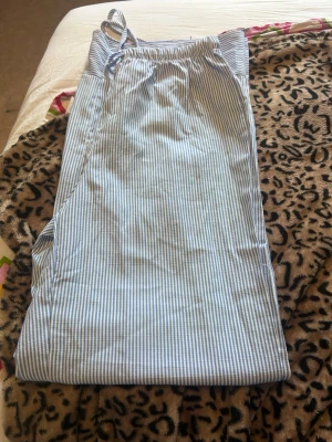 Randiga blåvita pyjamasbyxor - Säljer ett par klassiska pyjamasbyxor med tunna blå och vita ränder. Byxorna har resår i midjan och knytband för extra komfort. Tillverkade i ett mjukt och lätt tyg som passar perfekt för mysiga kvällar hemma.