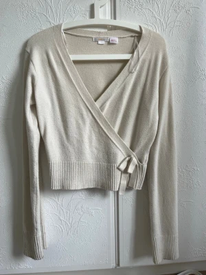 Beige omlottkofta från Gina Tricot - Snygg beige omlottkofta från Gina Tricot med långa ärmar och knytband i midjan. Koftan har ribbade muddar vid ärmslut och nederkant, och är tillverkad i ett mjukt stickat material. Perfekt för lager på lager och enkel att styla.