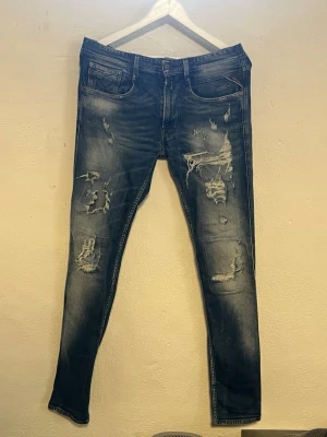 Replay anbass Jeans  - Snygga mörkblå slim jeans med slitningar och trasiga detaljer på både fram- och baksida. Jeansen har klassisk femficksdesign och är tillverkade i denim med stretch för en bekväm passform. Perfekta för en trendig och avslappnad stil.