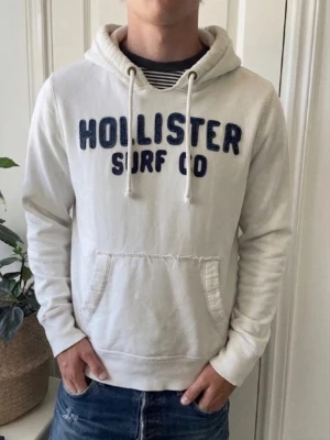 Vit hoodie från Hollister - Vit hoodie från Hollister med mörkblå broderad text 'HOLLISTER SURF CO' på bröstet. Klassisk känguruficka framtill och justerbar huva med vita snören. Mjuk bomullsmix som känns skön mot huden. Perfekt för en avslappnad och cool stil.