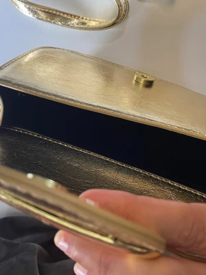 Guldig kuvertväska med axelrem - Snygg kuvertväska i metallic guld med matchande axelrem. Väskan har en stilren, rektangulär form och stängs med magnetknapp. Materialet är glansigt och ger en lyxig känsla. Perfekt för att lyfta vilken outfit som helst.