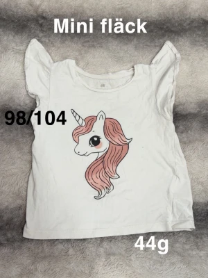 Vit t-shirt med enhörningstryck - Söt vit t-shirt med volangärmar och ett stort tryck av en enhörning med rosa man på framsidan. Perfekt för dig som gillar gulliga motiv och vill ha en bekväm och lekfull stil.🌸Mini fläck. Storlek 98/104