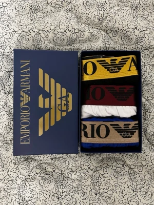  kalsonger 3-pack - kalsonger i olika färger: gul, vinröd, vit och blå. Alla har bred resår med Emporio Armani-logga och text. Snygg design och bekväm passform, perfekt för dig som vill ha stil även på underklädern.