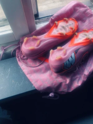 Nike Air Zoom rosa fotbollsskor - Säljer ett par Nike Air Zoom fotbollsskor i rosa med orange detaljer och svarta loggor. Skorna har dobbar för spel på gräs och är tillverkade i ett lätt syntetmaterial för bra grepp och snabbhet på planen. Snygg design och riktigt sköna att spela i Elite och originala priset är 3100kr helt nya testa dom engång.