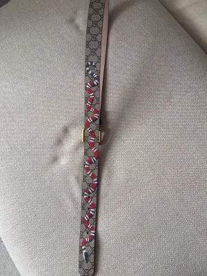 Gucci snake bälte äkta - Snyggt Gucci bälte med klassiskt GG-mönster och färgglad orm i rött, vitt och svart längs hela bandet. Bältet har ett guldfärgat spänne och är gjort i slitstarkt material med exklusiv känsla. Perfekt accessoar för att lyfta din outfit.