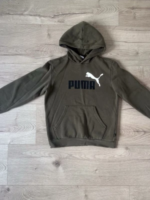 Olivgrön hoodie från Puma - Snygg olivgrön hoodie från Puma med stor vit logga och text på bröstet. Klassisk känguruficka framtill och ribbade muddar vid ärmar och nederkant. Perfekt för en avslappnad och sportig stil. Tillverkad i mjukt bomullsmaterial.