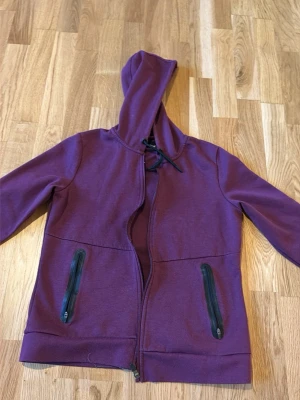 Lila hoodie från Crivit med dragkedja - Säljer en lila hoodie från Crivit med dragkedja framtill och två fickor med svarta dragkedjor. Tröjan har huva med snörning och är tillverkad i mjukt material, perfekt för chill dagar eller träning.