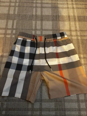 Rutig shorts med snörning - Säljer ett par snygga shorts med klassiskt rutigt mönster i beige, svart, vitt och rött. Shortsen har elastisk midja med svart snörning och en bakficka. Perfekta för sommaren och riktigt trendiga.