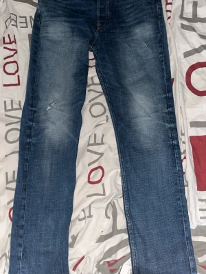 Blå raka jeans med slitningar - Säljer ett par klassiska blå jeans med raka ben och snygga slitningar på låret. Jeansen har en tvättad look och är tillverkade i denim. Perfekta för en avslappnad och trendig stil. Storleken står på andra bilden 