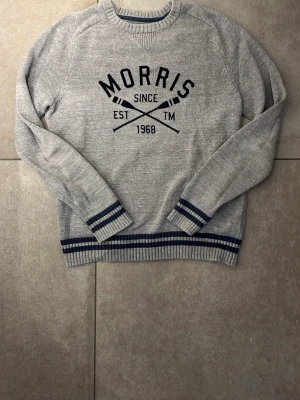Grå stickad tröja från Morris - Snygg grå stickad tröja från Morris med mörkblå text och detaljer. Tröjan har rund halsringning, ribbstickade muddar och nederkant med blå ränder. Perfekt för en avslappnad och stilren look.