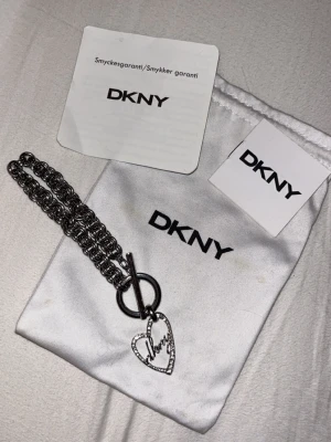 DKNY armband med hjärthänge  - Snyggt armband från DKNY i silverfärgad metall med ett grovt kedjemönster. Armbandet har ett hjärtformat hänge med gnistrande stenar och DKNY-logga i skrivstil. Låset är en dekorativ T-bar som ger en trendig touch.