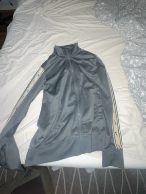 Grå Nike träningsjacka med beige detaljer - Snygg grå Nike träningsjacka med dragkedja och hög krage. Ärmarna har beige ränder med svarta loggor längs sidorna. Jackan har ribbade muddar och två fickor framtill. Perfekt för sport eller chill.