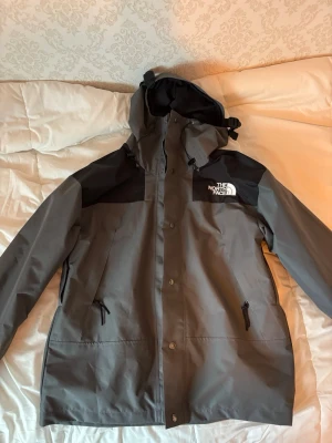 Grå vindjacka från The North Face - Snygg grå vindjacka från The North Face med svarta axelpartier och huva. Jackan har dragkedja och tryckknappar framtill, två sidofickor med dragkedja och justerbar huva. Perfekt för blåsiga dagar och outdoor-äventyr. Jättebra skick!!